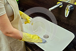 A woman is cleaning the bathroom by wiping the sink with a rag. Disinfection with yellow rubber gloves in the washroom