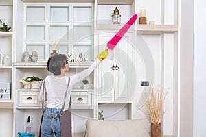Woman cleaning dust from bookshelf. Young girl sweeping shelf in living room. back view