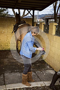 Woman cleaning her riding boots with her brown horse in the background