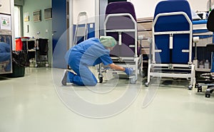Woman from the cleaning service of a hospital crouching cleaning the wheels of a patient chair in the operating room beforehand.