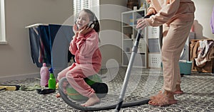 Woman cleans bathroom laundry room vacuums floor with vacuum cleaner. On equipment sits girl who accompanies mother in