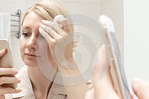 Woman cleansing her face with a cotton pad and watching a smartphone in her hand. Internet addiction concept