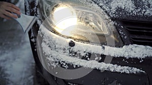 A woman clears snow from her car's headlights on a frosty day