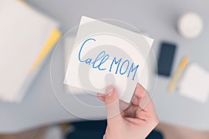 Woman clerk is sitting at office table holding note sticker with message `call mom`