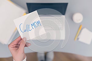 Woman clerk is sitting at office table holding note sticker with message `call mom`