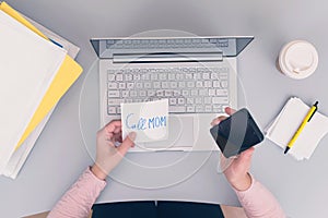 Woman clerk is sitting at office table holding note sticker with message `call mom`