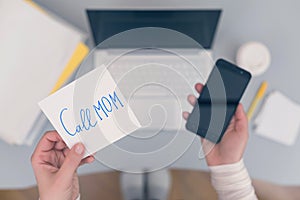 Woman clerk is sitting at office table holding note sticker with message `call mom`