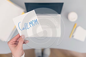 Woman clerk is sitting at office table holding note sticker with message `call mom`