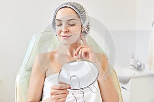Woman client of cosmetology salon satisfied with result of cosmetic procedure. Female looking at mirror and examining skin after
