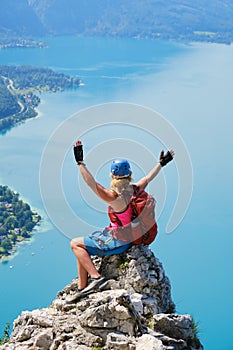 Woman with climbing helmet and via ferrata gear sits on top of a rock at Attersee, Austria, and spreads her arms. Happiness.
