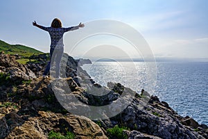 Woman climbing a stone with open arms and looking at the sea from the cliffs