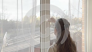 A woman closes a wide-open window made of PVC plastic and pulls the curtains.