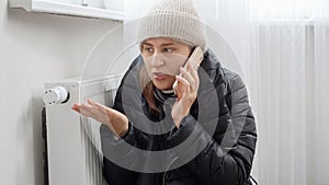 Woman in coat and hat at home calling repairman to fix heating system at house. Concept of energy crisis, high bills, broken