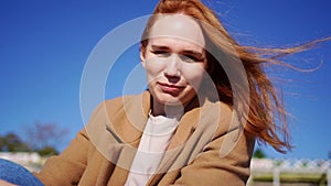 Woman in coat squints from sun and straightens hair, which is confused by wind.