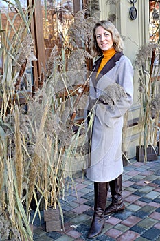 A woman in a coat stands near an autumn composition of dried flowers