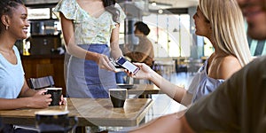 Woman In Coffee Shop Meeting Friend Paying Bill With Contactless Mobile Phone Payment