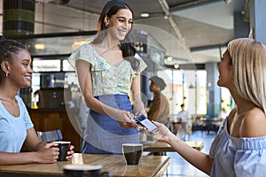 Woman In Coffee Shop Meeting Friend Paying Bill With Contactless Mobile Phone Payment