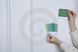 Woman with color sample cards choosing paint shade near white wall, closeup. Interior design