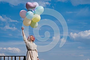 A woman with colored hair with an armful of balloons against a blue sky.