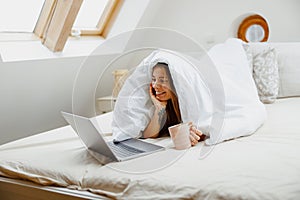 Woman in comfortable linens, happy on bed with laptop and coffee