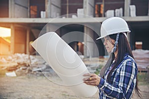 Woman construction engineer hold blueprint wear plaid shirt safety white hard hat at construction site industry labor worker.