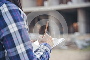 Woman construction engineer writing note wear plaid shirt safety white hard hat at construction site industry labor worker.