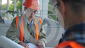 A woman in a construction helmet and protective vest with a drawing of the building sits opposite a male engineer