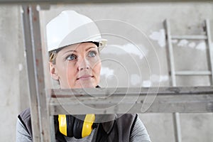Woman construction worker builder on ladder wearing white helmet and hearing protection headphones on interior site building