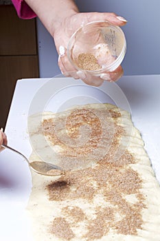 A woman is cooking buns with cinnamon. She rolls the stuffing onto the rolled dough. Mixed sugar and cinnamon.