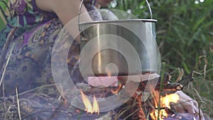 Woman cooking on fire in the forest. Cauldron, pot for cooking hanging on sticks over the fire. She is laying firewood under touri