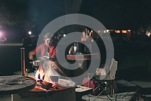 Woman cooking with fire wood and braai equipment by night. Tent and chairs in the foreground. Adventures in african national parks