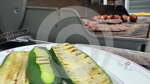 a woman cooking fresh grilled zucchini on a gas grill and then placed on a white serving plate with a silver metal spatula