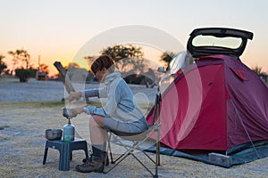 Woman cooking with gas stove in camping site at dusk. Gas burner, pot and smoke from boiling water, tent in the background. Advent