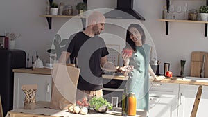 A woman cooks, the man brings the paper bags of food and puts vegetables and fruits from a paper bag on the table.