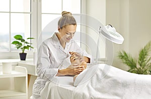 Woman cosmetologist checking client skin condition under lamp in beauty salon.