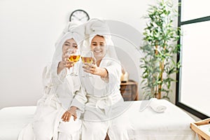 Woman couple wearing bathrobe toasting with champagne sitting on massage board at beauty center photo