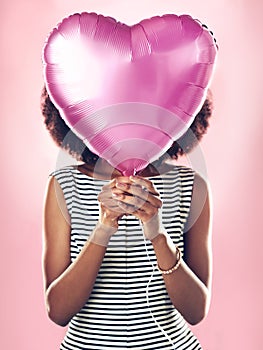 Woman cover face with heart balloon in studio, pink background and celebrate surprise birthday party. Female model hide