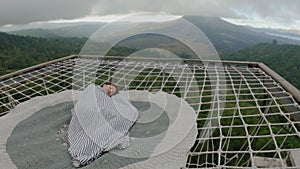 Woman covered with blanket sleeps on couch made of hammock net in open air above green misty abyss. Korean tourist is