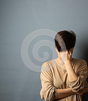 A woman covers her face with her hand, stands against the wall. Woman portrait on a gray background