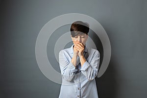 A woman covers her face with her hand, stands against the wall. Woman portrait on a gray background