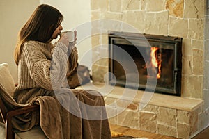 Woman in cozy sweater drinking cup of warm tea at fireplace in rustic room. Heating house in winter with wood burning stove. Young