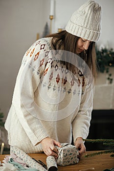 Woman in cozy sweater and hat wrapping christmas gift in paper on wooden table with festive decorations in decorated scandinavian