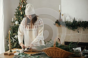 Woman in cozy sweater and hat wrapping christmas gift in paper on wooden table with festive decorations in decorated scandinavian