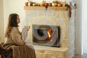 Woman in cozy sweater holding cup of warm tea at fireplace, autumn hygge. Heating house with wood burning stove. Young stylish