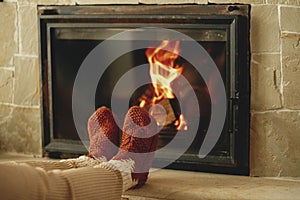 Woman in cozy wool socks warming up feet at fireplace in rustic room. Heating house in winter with wood burning stove. Young