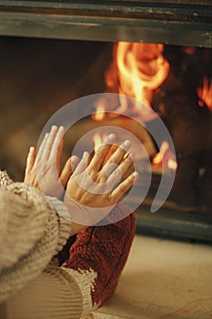 Woman in cozy wool socks warming up feet and hands at fireplace in rustic room. Heating house in winter with wood burning stove.