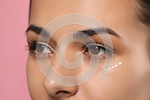 Woman with cream around eye on pink background, closeup. Skin care