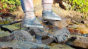 A woman crosses the river on the stones. Female legs close up in sneakers that chlnm on wet stones crossing a small