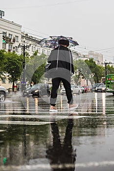 A woman crossing a street and a zebra with an umbrella in the rain.