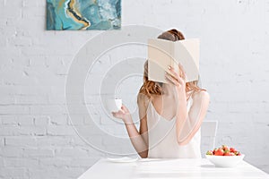 woman with cup of coffee and bowl of strawberries covering face with book